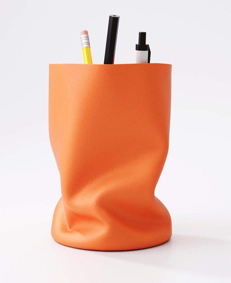 May include: An orange desk organizer with a unique, crumpled design. The container holds a yellow pencil, a black pencil, and a black and white pen. The organizer is made of a smooth material and sits on a white surface.