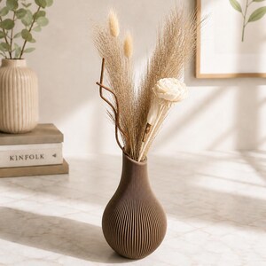May include: A brown, ribbed vase holds dried ornamental grasses and a white flower. The vase has a teardrop shape and sits on a marble surface. A second beige vase with eucalyptus is in the background.