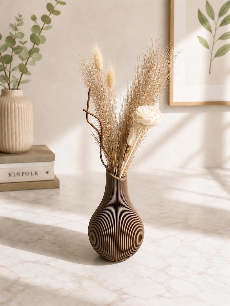 May include: A brown, ribbed vase holds dried ornamental grasses and a white flower. The vase has a teardrop shape and sits on a marble surface. A second beige vase with eucalyptus is in the background.