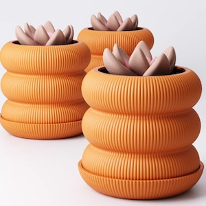 May include: Three orange ribbed ceramic plant pots, each containing a faux succulent with pink and green leaves. The pots have a unique stacked design with a matching saucer base. The background is plain white.