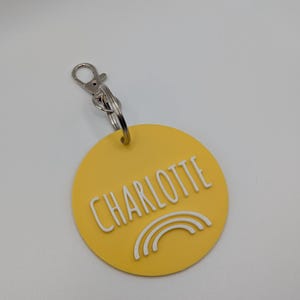 May include: A yellow circular keychain with the name "CHARLOTTE" in white capital letters. Below the name is a white rainbow design. The keychain has a silver clasp and ring.