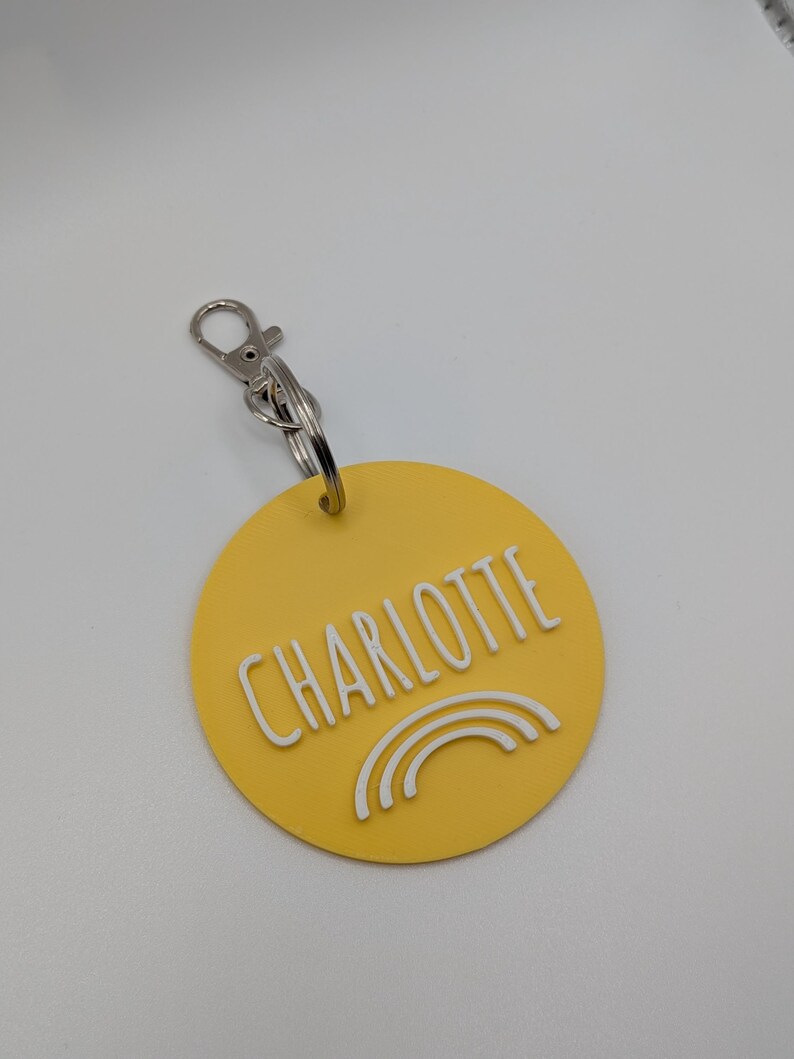May include: A yellow circular keychain with the name "CHARLOTTE" in white capital letters. Below the name is a white rainbow design. The keychain has a silver clasp and ring.