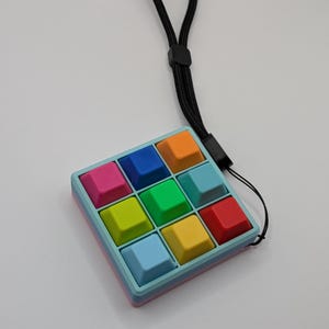 May include: A colorful, handheld fidget toy with nine square buttons in various colors, including pink, blue, orange, green, yellow, and red. The toy has a light blue frame and a black lanyard for easy carrying.