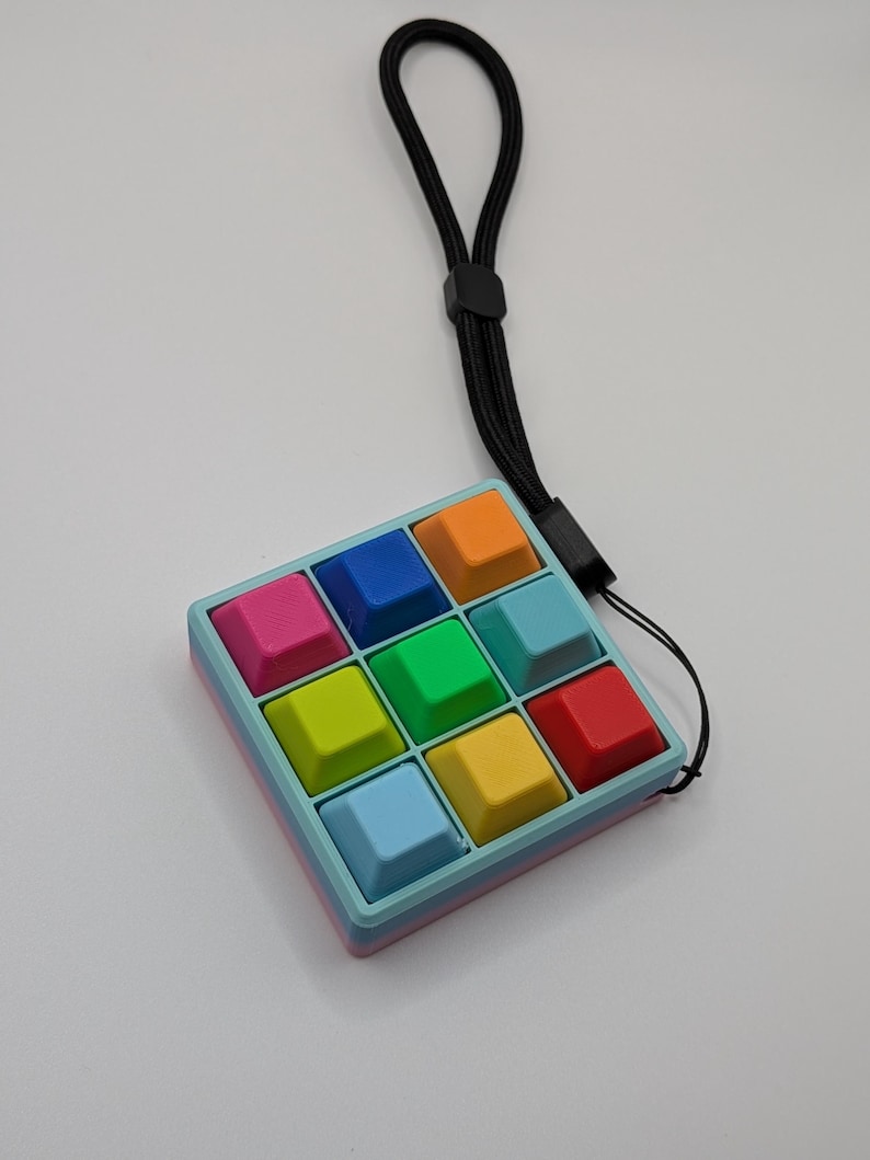 May include: A colorful, handheld fidget toy with nine square buttons in various colors, including pink, blue, orange, green, yellow, and red. The toy has a light blue frame and a black lanyard for easy carrying.