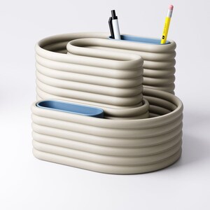 May include: A beige desk organiser with a wavy design, featuring three compartments with light blue interiors. Two pens and a yellow pencil are visible. The organiser is made of a ridged material, suitable for office or home use.