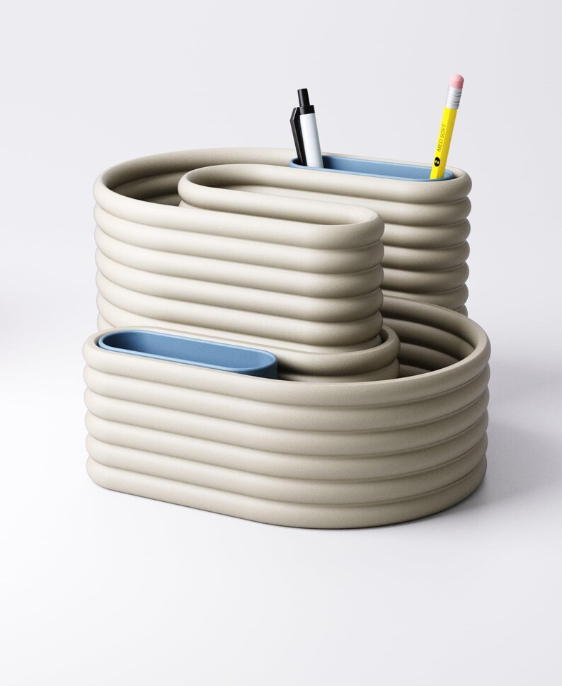 May include: A beige desk organiser with a wavy design, featuring three compartments with light blue interiors. Two pens and a yellow pencil are visible. The organiser is made of a ridged material, suitable for office or home use.