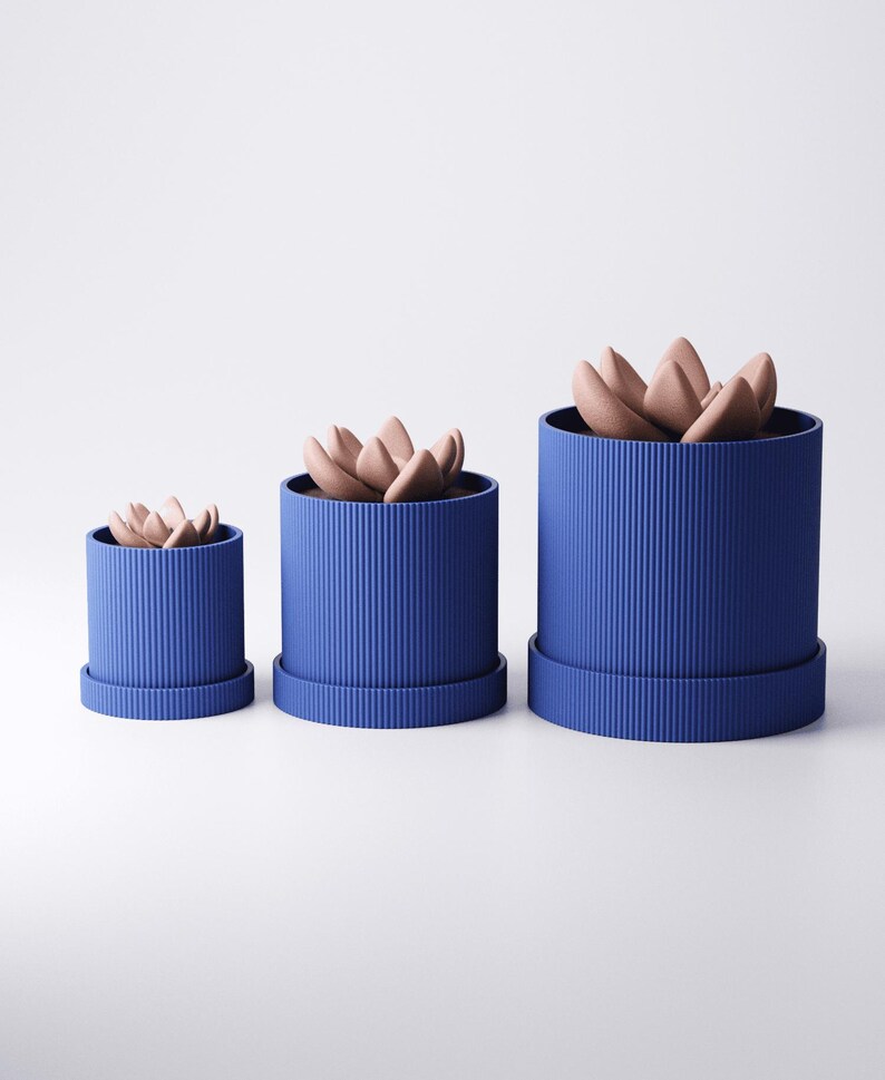 May include: Three blue cylindrical planters of varying sizes, each with a ribbed texture and a matching saucer base. The planters contain faux succulents with light brown, petal-like leaves. The arrangement is set against a white background.