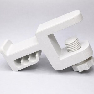 Puede incluir: Una abrazadera blanca en forma de C con un mecanismo de tornillo para ajustar el agarre. La abrazadera tiene una base rectangular con tres agujeros y un brazo superior. El tornillo est&aacute; situado en el lateral y el mango tiene forma de estrella.