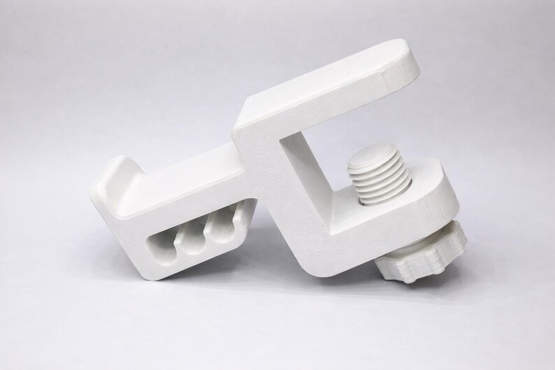 Puede incluir: Una abrazadera blanca en forma de C con un mecanismo de tornillo para ajustar el agarre. La abrazadera tiene una base rectangular con tres agujeros y un brazo superior. El tornillo est&aacute; situado en el lateral y el mango tiene forma de estrella.