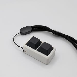 May include: A white rectangular device with two black square buttons. It has a black lanyard attached. The device is designed to be worn around the neck or wrist. The background is plain white.
