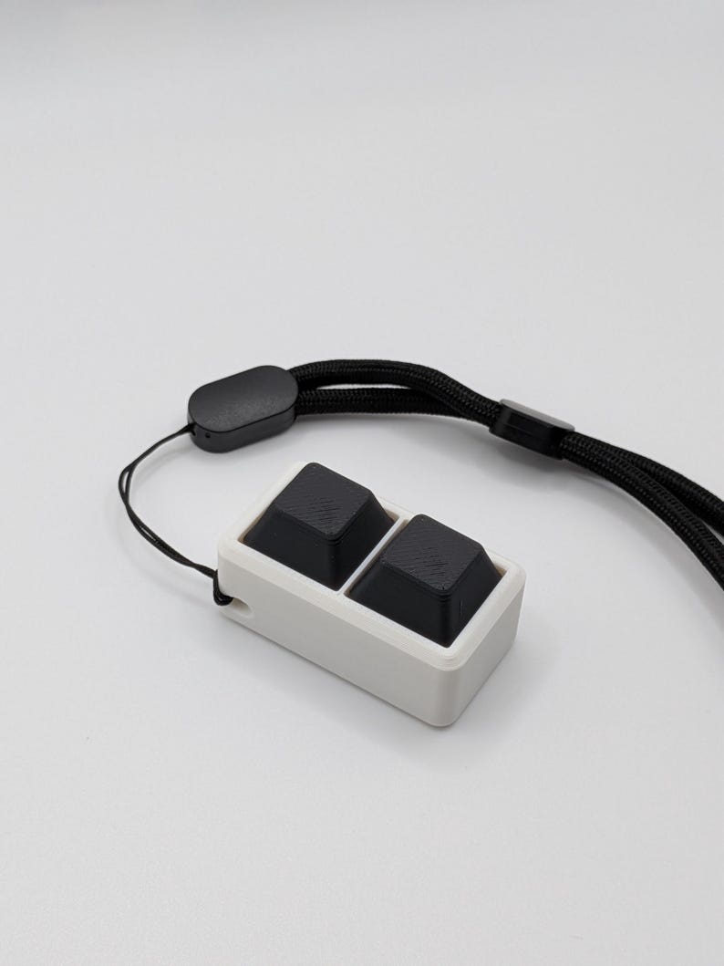May include: A white rectangular device with two black square buttons. It has a black lanyard attached. The device is designed to be worn around the neck or wrist. The background is plain white.
