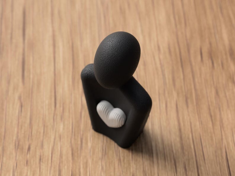 Puede incluir: Una escultura abstracta negra con cabeza y cuerpo texturizados, con un detalle blanco en forma de coraz&oacute;n en la parte delantera. La escultura est&aacute; sobre un fondo de madera.