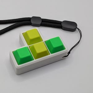 May include: A white pendant featuring four colorful keyboard keys in a Tetris-like shape. The keys are green and yellow, set in a white frame, and attached to a black lanyard. The pendant is designed to be worn.