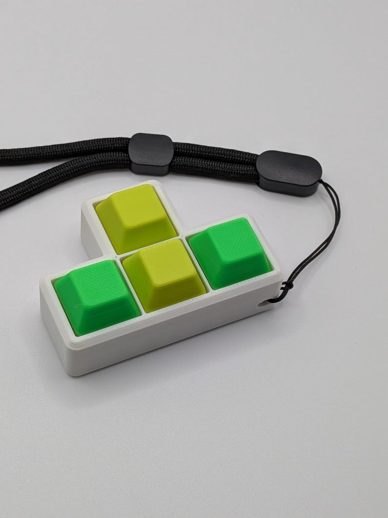 May include: A white pendant featuring four colorful keyboard keys in a Tetris-like shape. The keys are green and yellow, set in a white frame, and attached to a black lanyard. The pendant is designed to be worn.