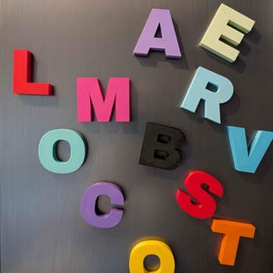 May include: Colorful magnetic alphabet letters in various colors, including red, pink, purple, and yellow, are arranged on a gray metallic surface. The letters are in a bold, block style, suitable for educational or decorative purposes.