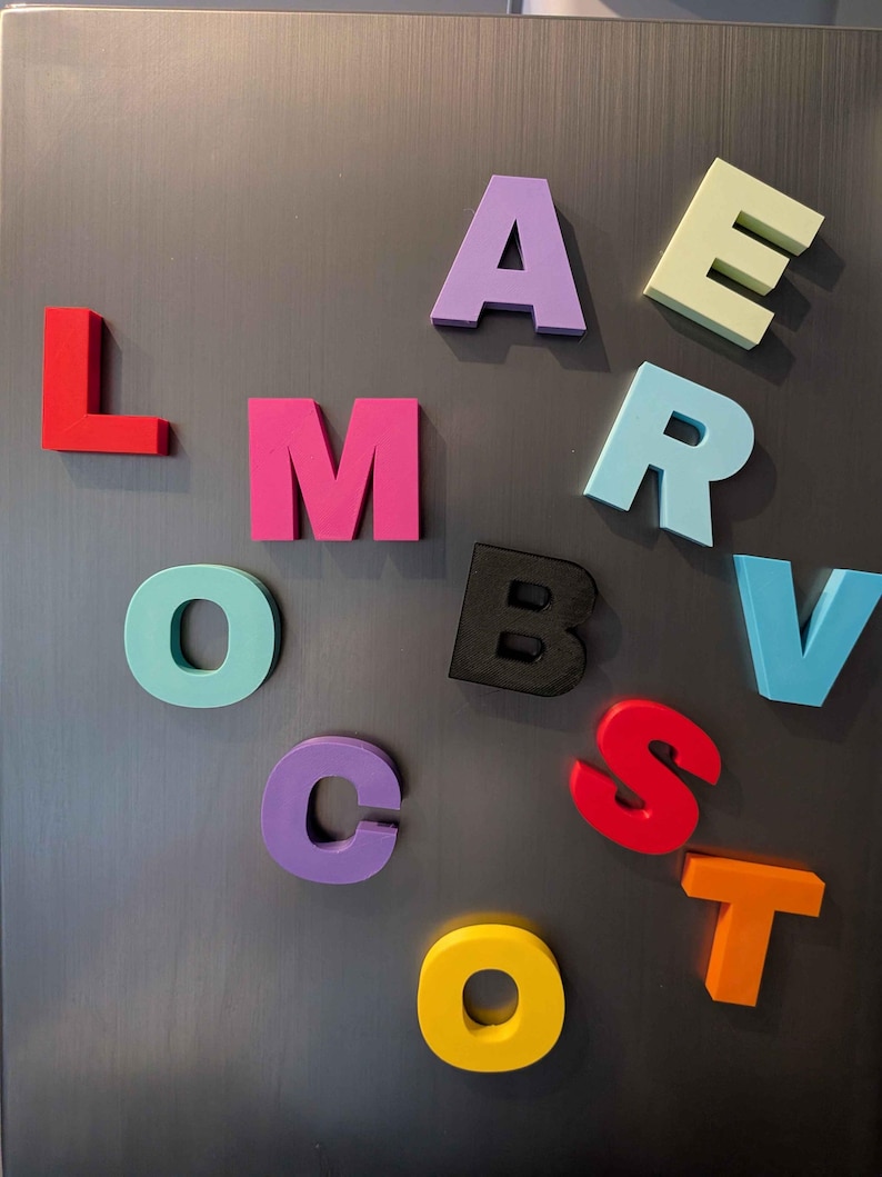 May include: Colorful magnetic alphabet letters in various colors, including red, pink, purple, and yellow, are arranged on a gray metallic surface. The letters are in a bold, block style, suitable for educational or decorative purposes.