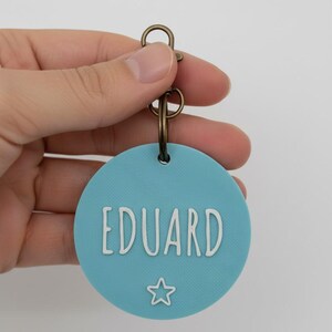 May include: A round, light blue pet tag with the name "EDUARD" in white, sans-serif font. A white star is below the name. The tag is attached to a bronze-colored split ring and clasp.