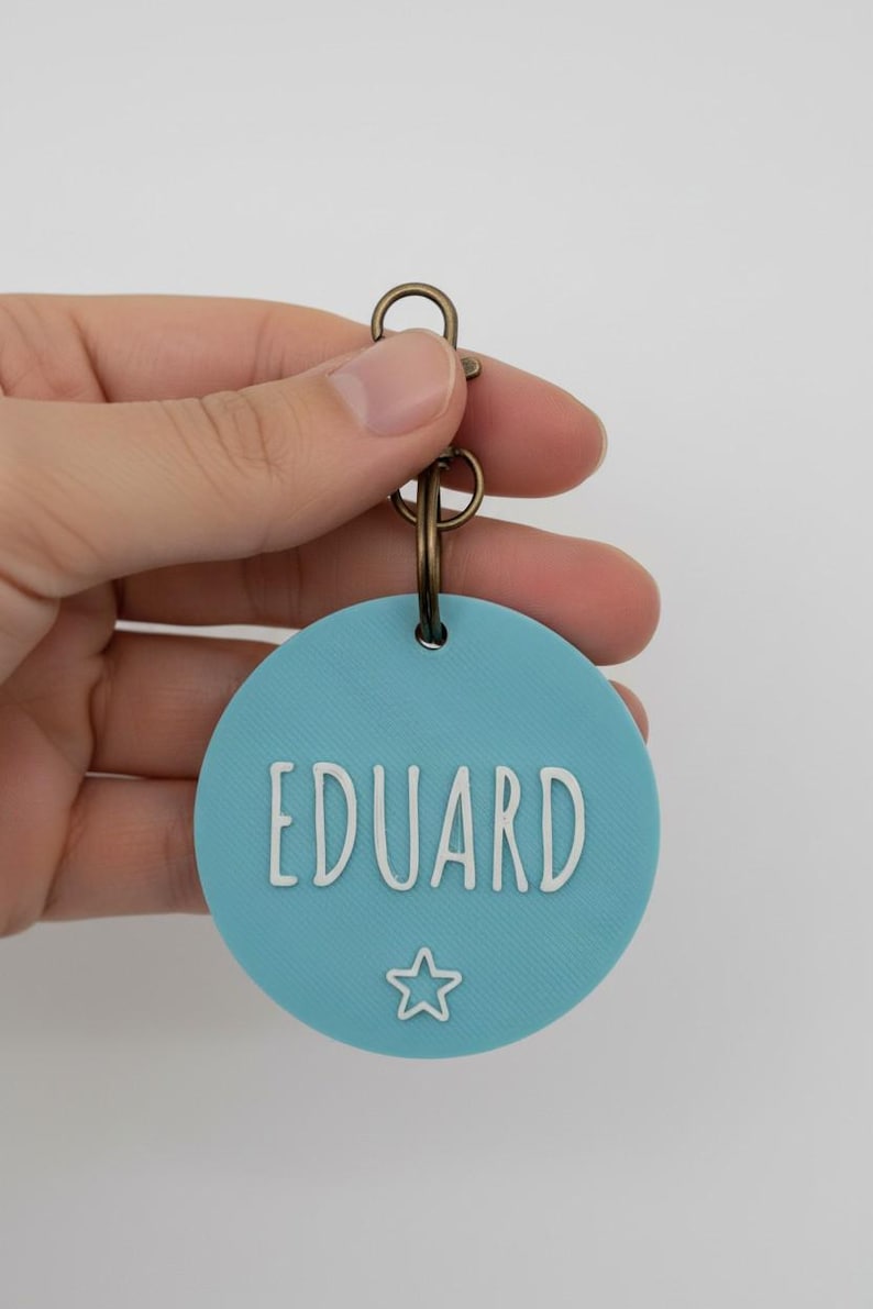 May include: A round, light blue pet tag with the name "EDUARD" in white, sans-serif font. A white star is below the name. The tag is attached to a bronze-colored split ring and clasp.