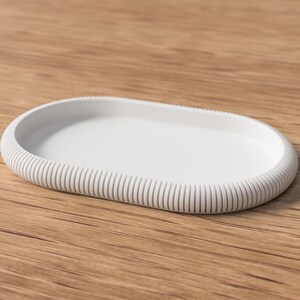 Puede incluir: Una bandeja blanca de forma ovalada con borde acanalado. La bandeja est&aacute; hecha de un material s&oacute;lido y se encuentra sobre una superficie de madera. El dise&ntilde;o es minimalista y moderno, adecuado para diversos usos.