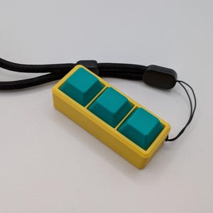 May include: A yellow rectangular keychain with three teal square buttons. The keychain is attached to a black lanyard. The buttons have a textured surface. The background is a solid white color.