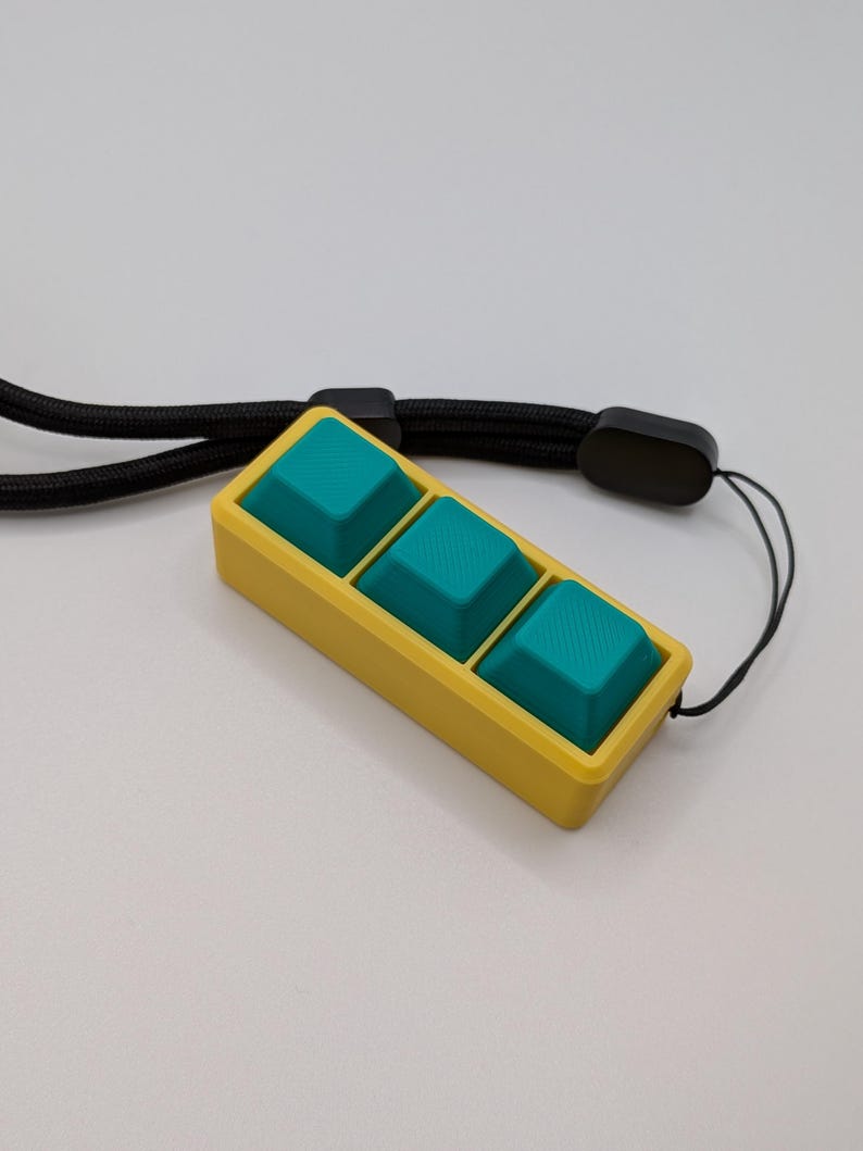 May include: A yellow rectangular keychain with three teal square buttons. The keychain is attached to a black lanyard. The buttons have a textured surface. The background is a solid white color.