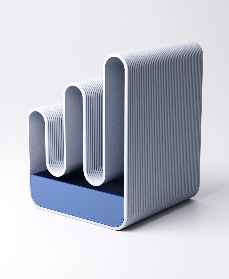 May include: A modern, abstract desk organizer in shades of gray and blue. The design features a series of curved, vertical elements with a ribbed texture, set above a rectangular blue base. The overall aesthetic is minimalist and contemporary.