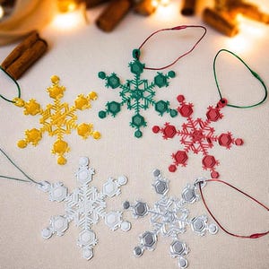 May include: A collection of snowflake ornaments in yellow, green, red, silver, and gray. Each ornament has a unique design and is suspended by a matching colored string. The ornaments are arranged on a neutral surface, with blurred lights and cinnamon sticks in the background.