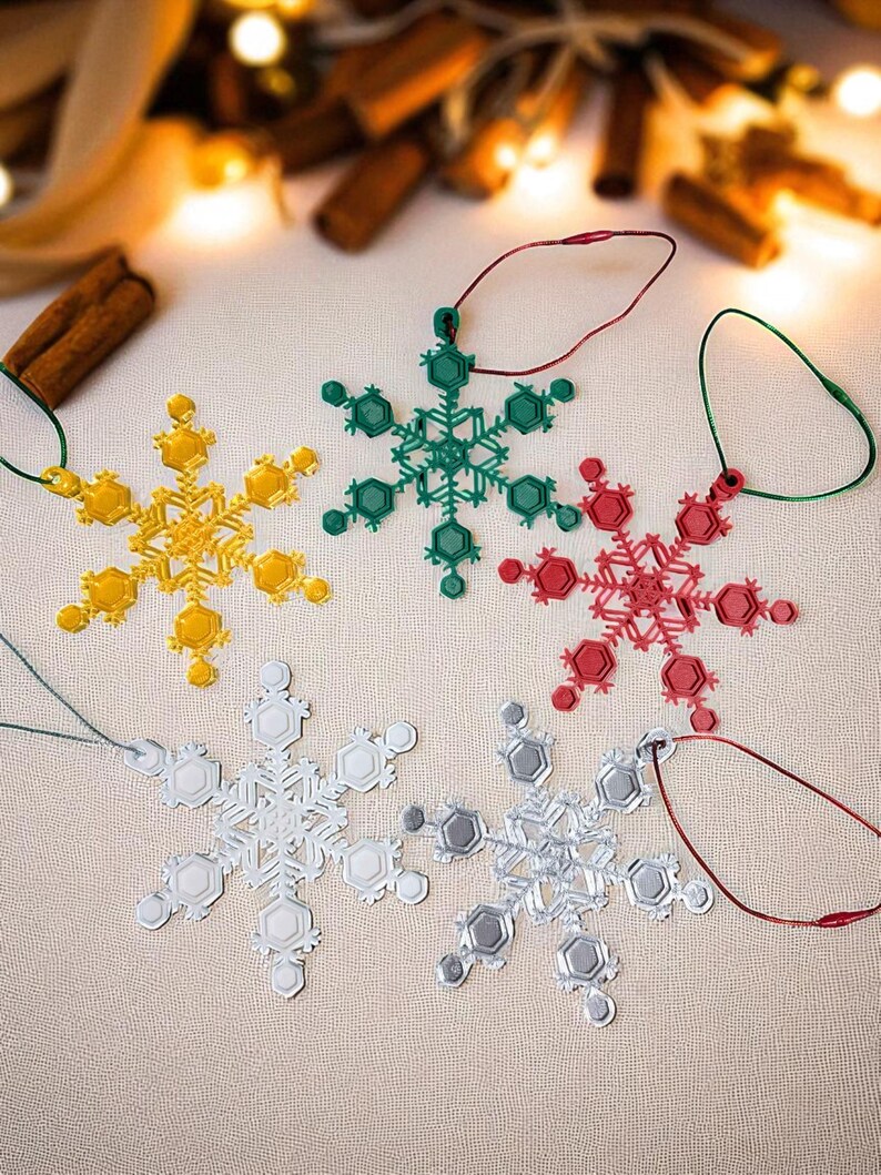 May include: A collection of snowflake ornaments in yellow, green, red, silver, and gray. Each ornament has a unique design and is suspended by a matching colored string. The ornaments are arranged on a neutral surface, with blurred lights and cinnamon sticks in the background.