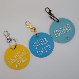 May include: Three personalized keychains with names and details. One yellow keychain says "CHARLOTTE" with a rainbow graphic. A blue keychain says "OLIVER CLASS 3B". The third blue keychain says "EDUARD" with a star graphic.