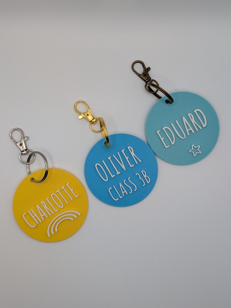 May include: Three personalized keychains with names and details. One yellow keychain says "CHARLOTTE" with a rainbow graphic. A blue keychain says "OLIVER CLASS 3B". The third blue keychain says "EDUARD" with a star graphic.
