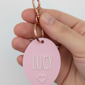 May include: A pink oval keychain with the name "LUCY" in white letters and a small heart symbol. The keychain has a rose gold clasp and ring.