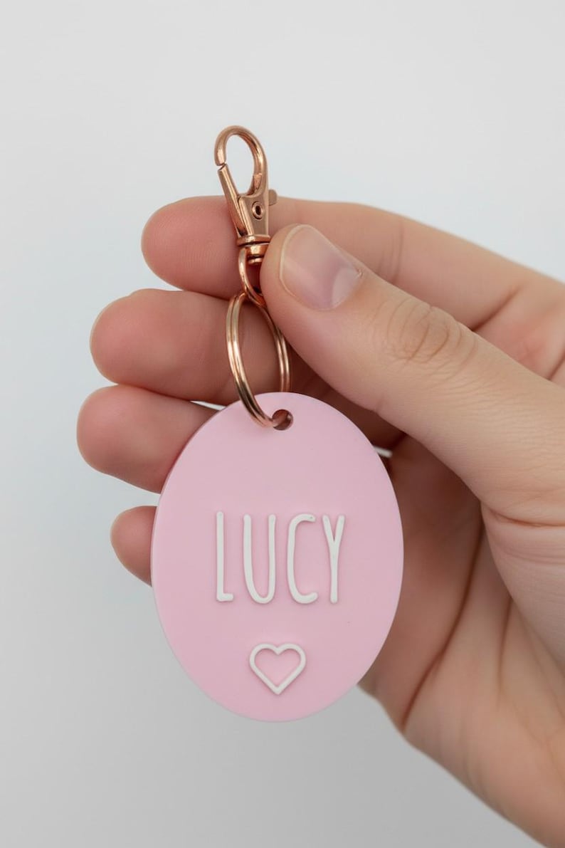 May include: A pink oval keychain with the name "LUCY" in white letters and a small heart symbol. The keychain has a rose gold clasp and ring.