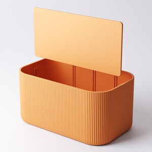 May include: An orange rectangular storage box with a lid slightly open. The box features vertical ribbed detailing on the sides and a smooth, flat lid. The box is made of a solid, matte material.