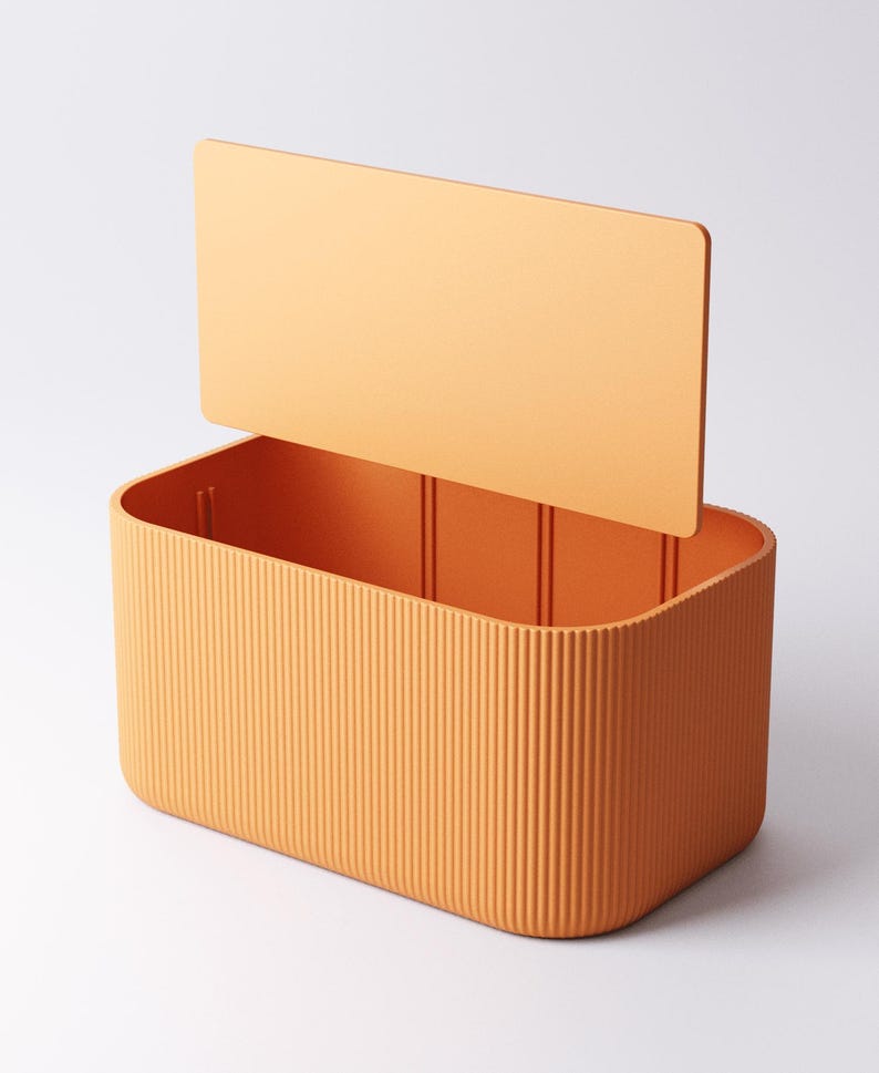 May include: An orange rectangular storage box with a lid slightly open. The box features vertical ribbed detailing on the sides and a smooth, flat lid. The box is made of a solid, matte material.