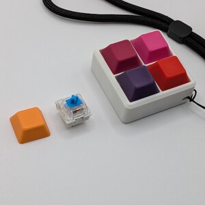 May include: A collection of colorful keyboard keycaps in a white tray, with a separate orange keycap and a clear switch. The keycaps are various shades of red, pink, and purple. A black lanyard is attached to the tray.