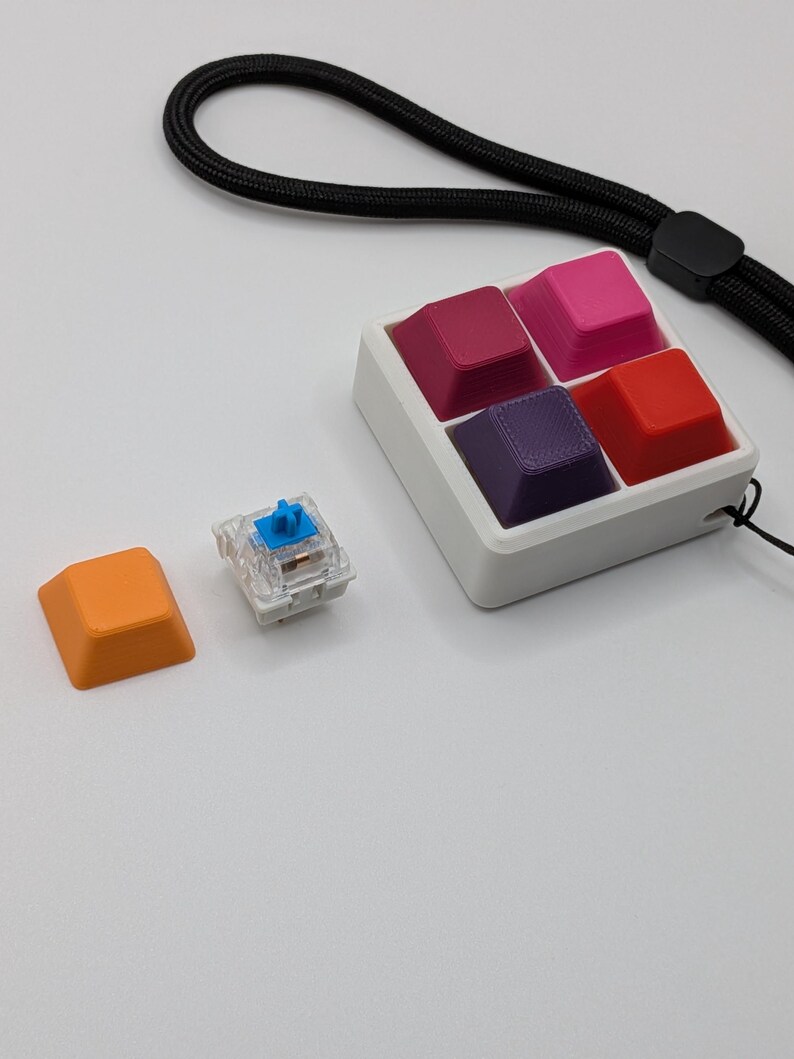 May include: A collection of colorful keyboard keycaps in a white tray, with a separate orange keycap and a clear switch. The keycaps are various shades of red, pink, and purple. A black lanyard is attached to the tray.