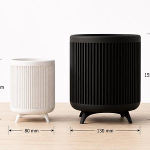 Puede incluir: Dos maceteros cil&iacute;ndricos, uno blanco y otro negro, con detalles acanalados verticales. El macetero blanco mide 9,5 cm de alto y 8 cm de ancho, mientras que el negro mide 15,5 cm de alto y 13 cm de ancho. Ambos tienen tres patas peque&ntilde;as.