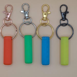 May include: Four keychains with different colored accents. The keychains have a metal clasp and ring, with a rectangular silicone accent in red, green, blue, and yellow. The metal hardware is in rose gold, silver, gold, and black.