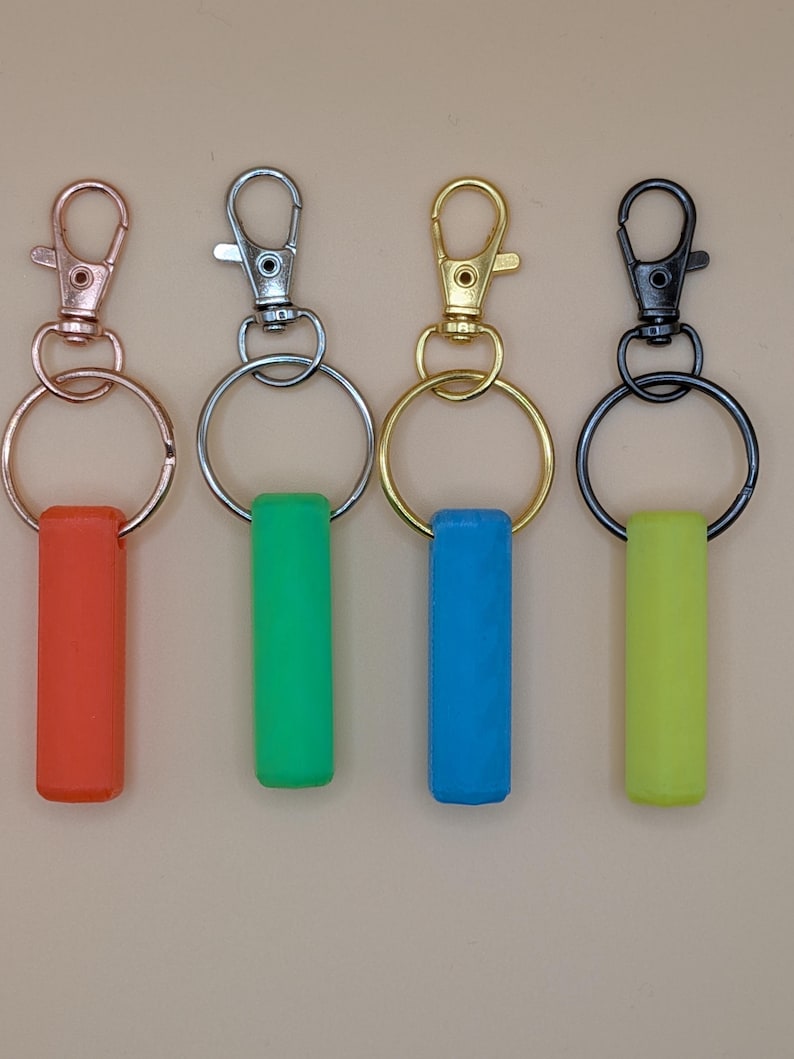May include: Four keychains with different colored accents. The keychains have a metal clasp and ring, with a rectangular silicone accent in red, green, blue, and yellow. The metal hardware is in rose gold, silver, gold, and black.