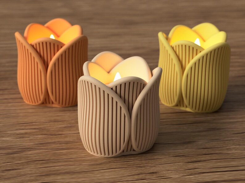 May include: Three tulip-shaped candle holders in shades of orange, beige, and yellow. Each holder has a vertical ribbed design and holds a lit candle, creating a warm glow. The candle holders are on a wooden surface.
