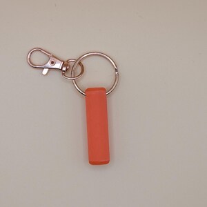 May include: A keychain with a coral-colored rectangular charm. The keychain has a silver-colored split ring and a lobster clasp. The background is a solid, light beige color.