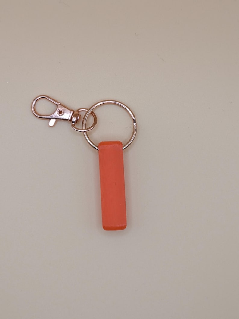 May include: A keychain with a coral-colored rectangular charm. The keychain has a silver-colored split ring and a lobster clasp. The background is a solid, light beige color.