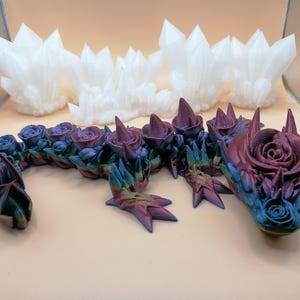 May include: A fantasy-themed sculpture featuring a dragon with a rose-like head and body. The dragon is painted in shades of blue, purple, and green. Several white crystal formations are in the background. The dragon's body is adorned with rose-shaped details.