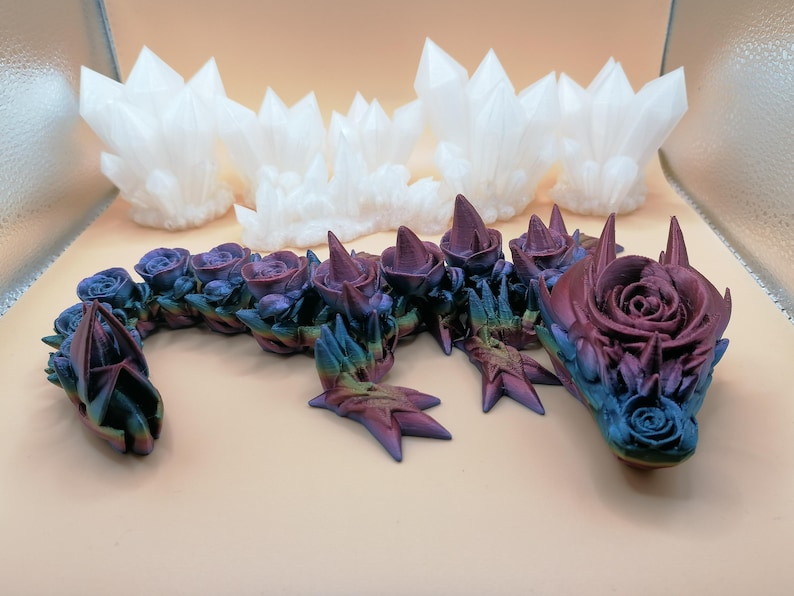 May include: A fantasy-themed sculpture featuring a dragon with a rose-like head and body. The dragon is painted in shades of blue, purple, and green. Several white crystal formations are in the background. The dragon's body is adorned with rose-shaped details.
