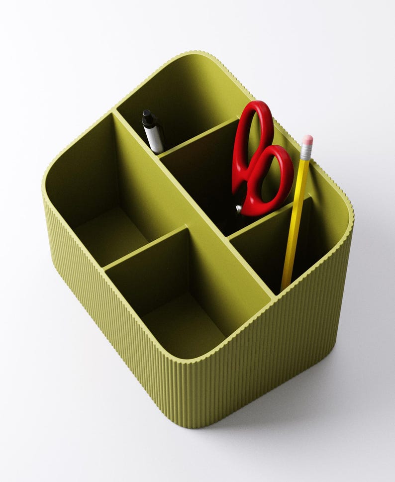 May include: A green desk organizer with multiple compartments. It contains a black pen, red scissors, and a yellow pencil with a pink eraser. The organizer has a ribbed exterior and is designed to hold office supplies.