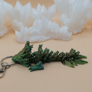May include: A green and bronze dragon keychain with a silver clasp and ring. The dragon is detailed with textured scales and spines. White crystal formations are in the background.