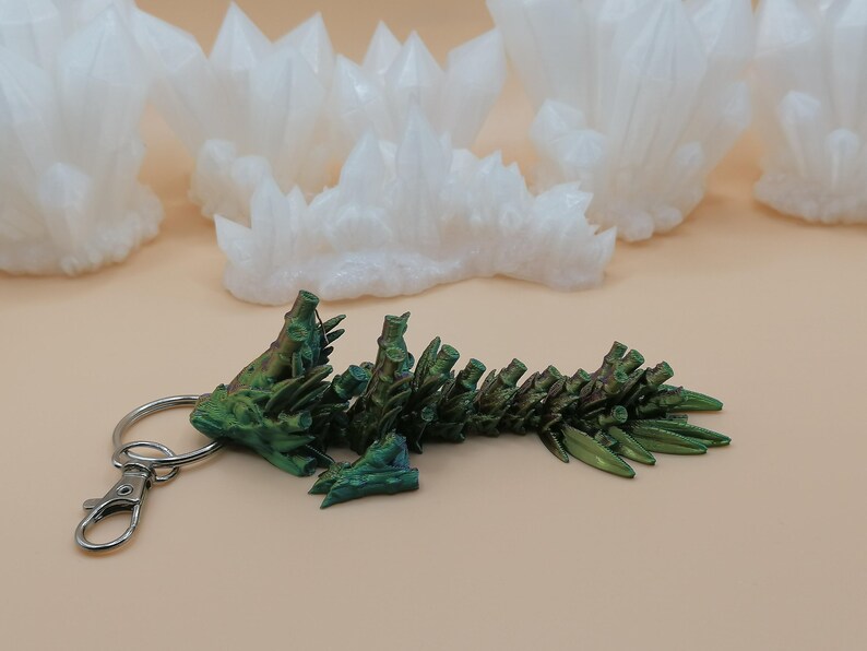 May include: A green and bronze dragon keychain with a silver clasp and ring. The dragon is detailed with textured scales and spines. White crystal formations are in the background.