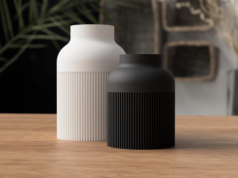 May include: Two modern ceramic vases. One is white, the other is black. Both have a cylindrical shape with a ribbed texture on the lower half. The vases are displayed on a wooden surface, with a blurred background of greenery and abstract art.