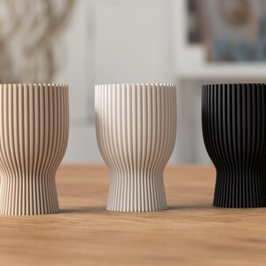 Pu&ograve; includere: Tre vasi decorativi in legno con scanalature verticali, nei colori beige, bianco sporco e nero. I vasi hanno una base arrotondata e una parte superiore pi&ugrave; ampia. Sono su una superficie di legno.