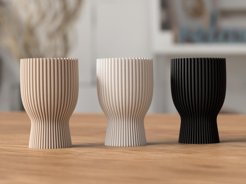 Pu&ograve; includere: Tre vasi decorativi in legno con scanalature verticali, nei colori beige, bianco sporco e nero. I vasi hanno una base arrotondata e una parte superiore pi&ugrave; ampia. Sono su una superficie di legno.